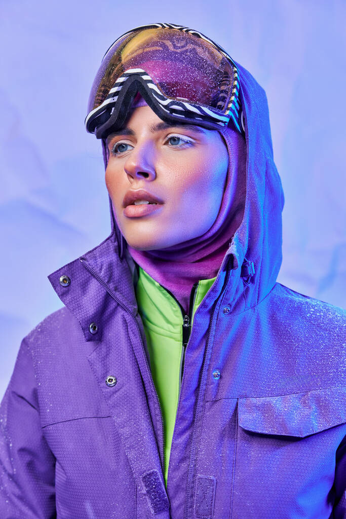 ropogós téli levegő, fiatal nő símaszkban, googles és meleg kabát néz el lila háttér - Fotó, kép