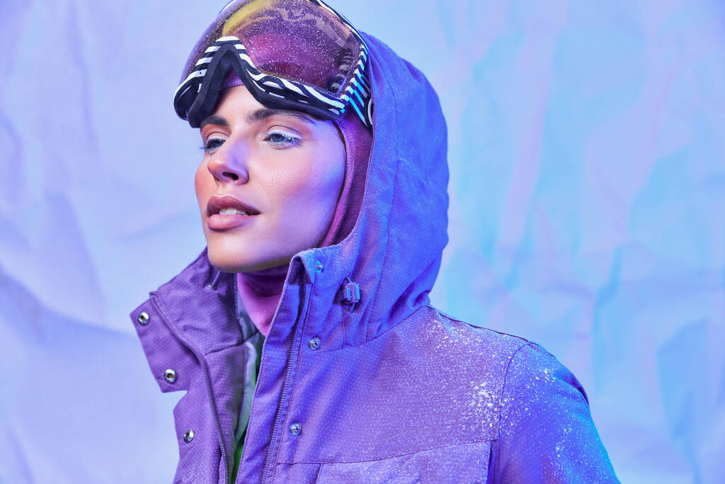 frosty winter air, young woman in ski mask, googles and warm jacket looking away on purple backdrop - Photo, Image