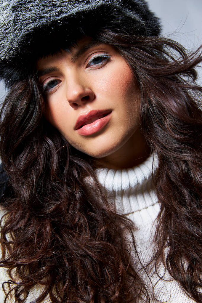 csinos nő műszőrme kalap és pulóver nézi kamera szürke háttér, téli szépség - Fotó, kép