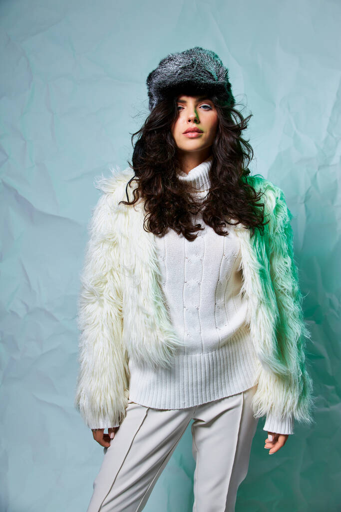стильная брюнетка в белом искусственном меховом пиджаке, зимней шляпе и свитере позирует на сером фоне - Фото, изображение