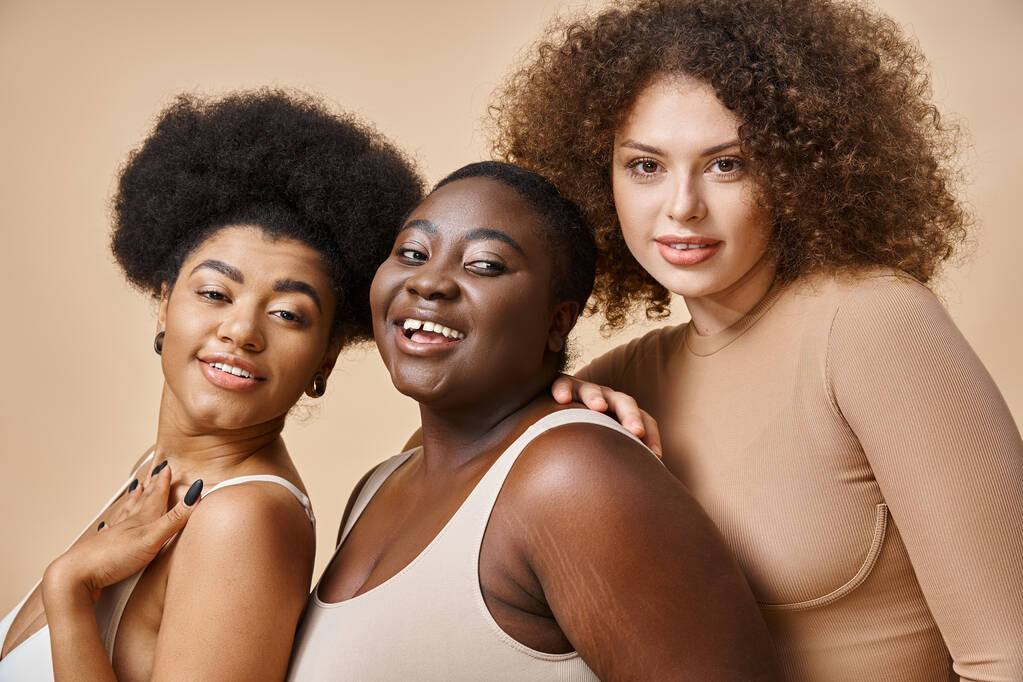vreugdevolle multi-etnische plus size vrouwen in ondergoed poseren op beige achtergrond, zelfvertrouwen en charme - Foto, afbeelding