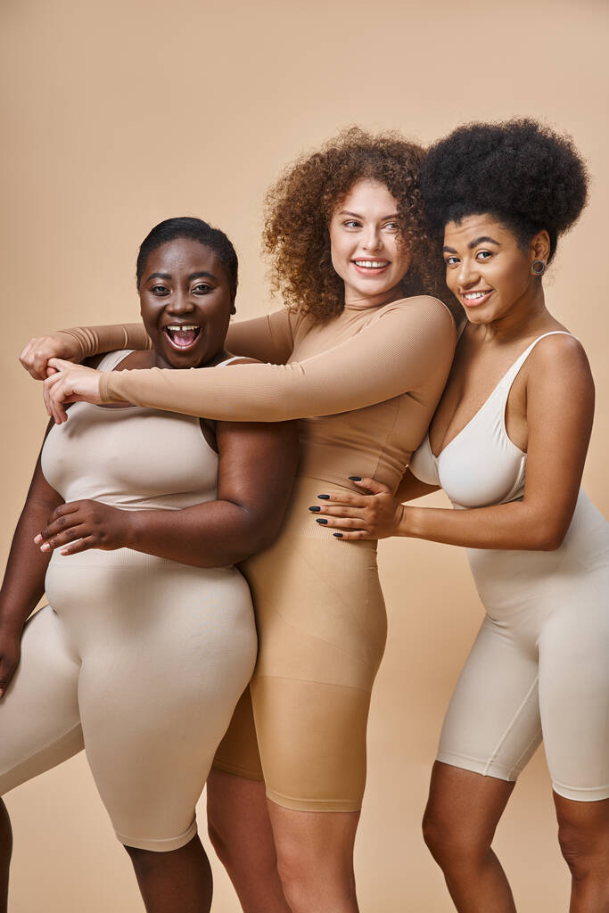 aufgeregte multirassische Plus-Size-Frauen in Dessous, die sich umarmen und über beige lachen, Körperpositivität - Foto, Bild