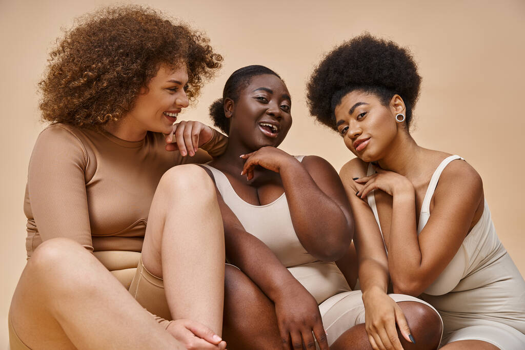 bezstarostný africký Američan plus velikost žena ve spodním prádle smích v blízkosti multietnické přítelkyně na béžové - Fotografie, Obrázek
