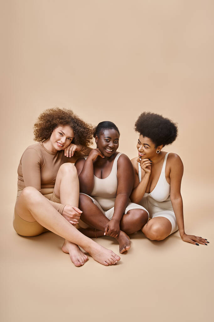 fröhliche multiethnische körperpositive Frauen in Dessous sitzen und lächeln auf beige, Selbstwertgefühl - Foto, Bild