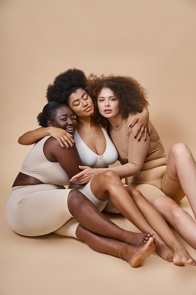 multiethnische Plus-Size-Freundinnen in Unterwäsche sitzen und umarmen auf grauer Zweisamkeit - Foto, Bild