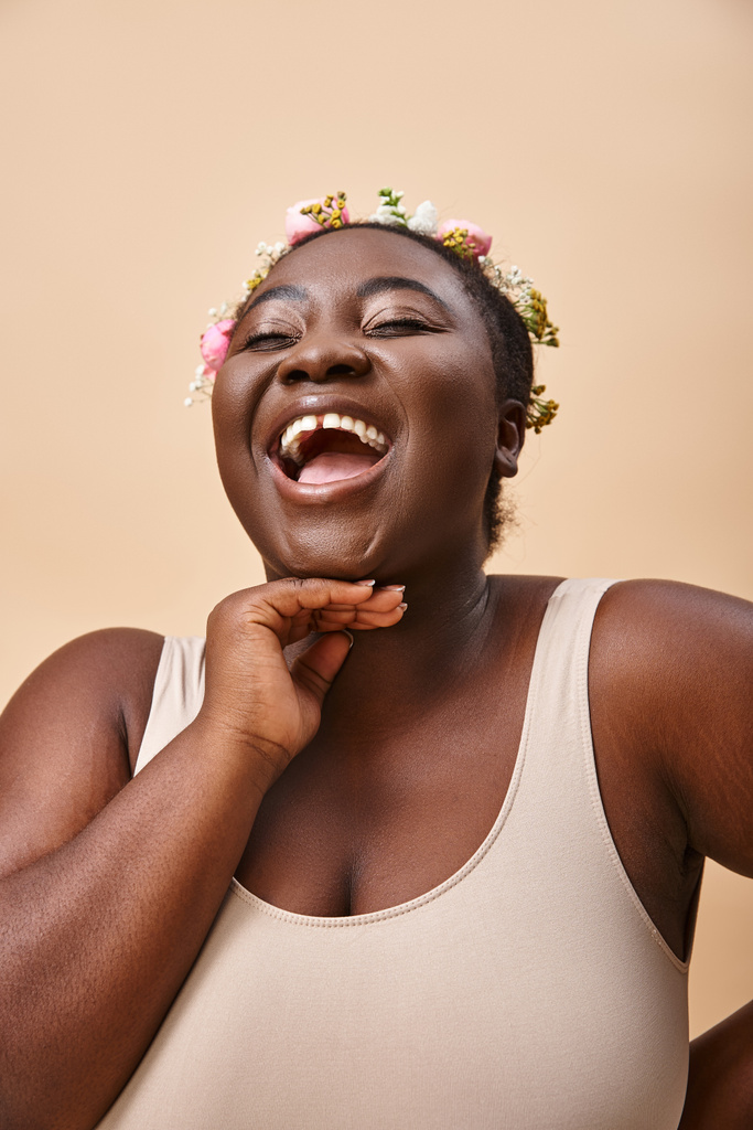 ενθουσιασμένοι συν μέγεθος αφροαμερικανή γυναίκα με λουλούδια στα μαλλιά γέλιο σε μπεζ, θετικότητα στο σώμα - Φωτογραφία, εικόνα