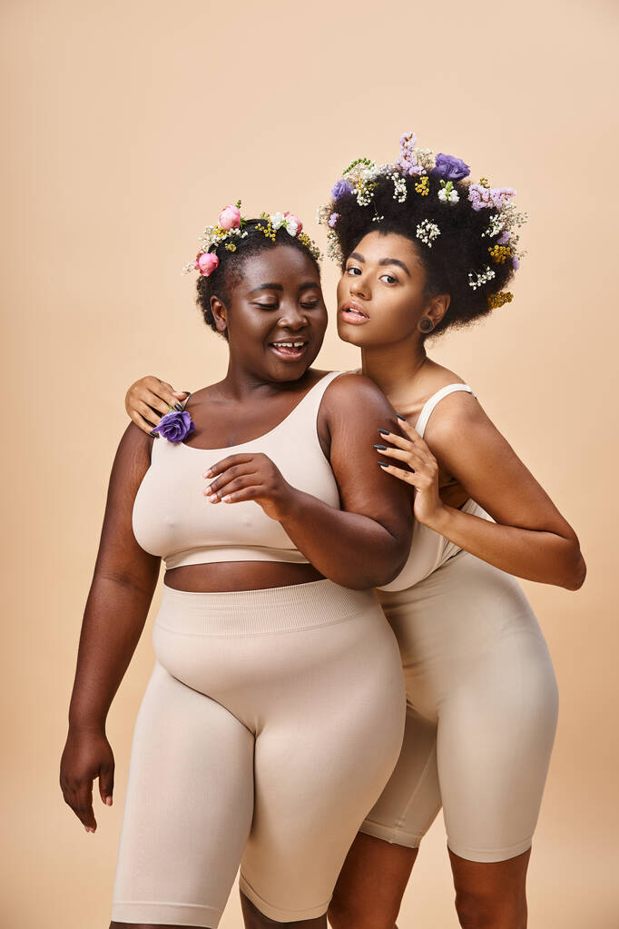 lächelnde afrikanisch-amerikanische Frauen mit Blumen im Haar posieren in Unterwäsche auf beige, plus size beauty - Foto, Bild