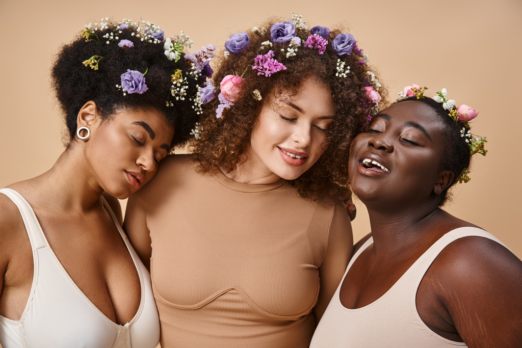 πολυφυλετικές γυναίκες συν μέγεθος με εσώρουχα με πολύχρωμα λουλούδια στα μαλλιά και κλειστά μάτια σε μπεζ - Φωτογραφία, εικόνα