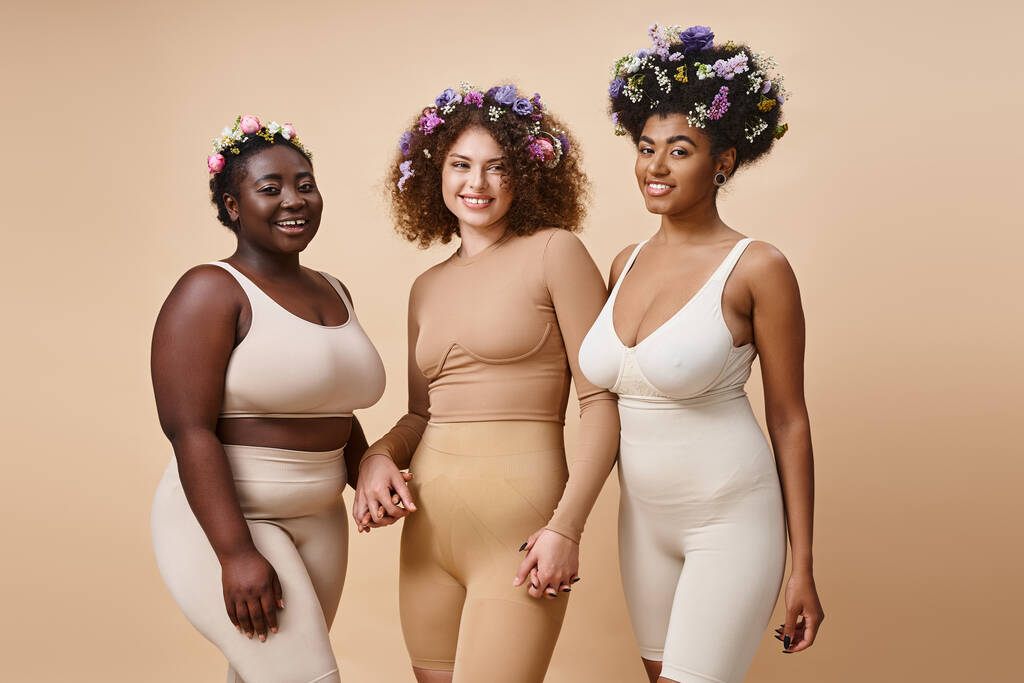 radostné multietnické přítelkyně ve spodním prádle s barevnými květy ve vlasech na béžové, plus velikost krásy - Fotografie, Obrázek