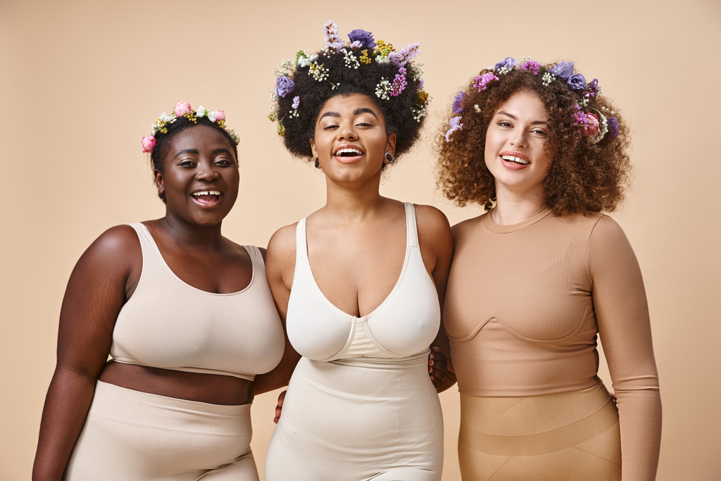 lächelnde multiethnische Freundinnen in Dessous mit bunten Blumen im Haar auf beige, plus size beauty - Foto, Bild