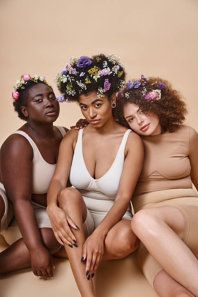 diverse multi-etnische vriendinnen in lingerie met bloemen in haar zitten op beige, plus size schoonheid - Foto, afbeelding