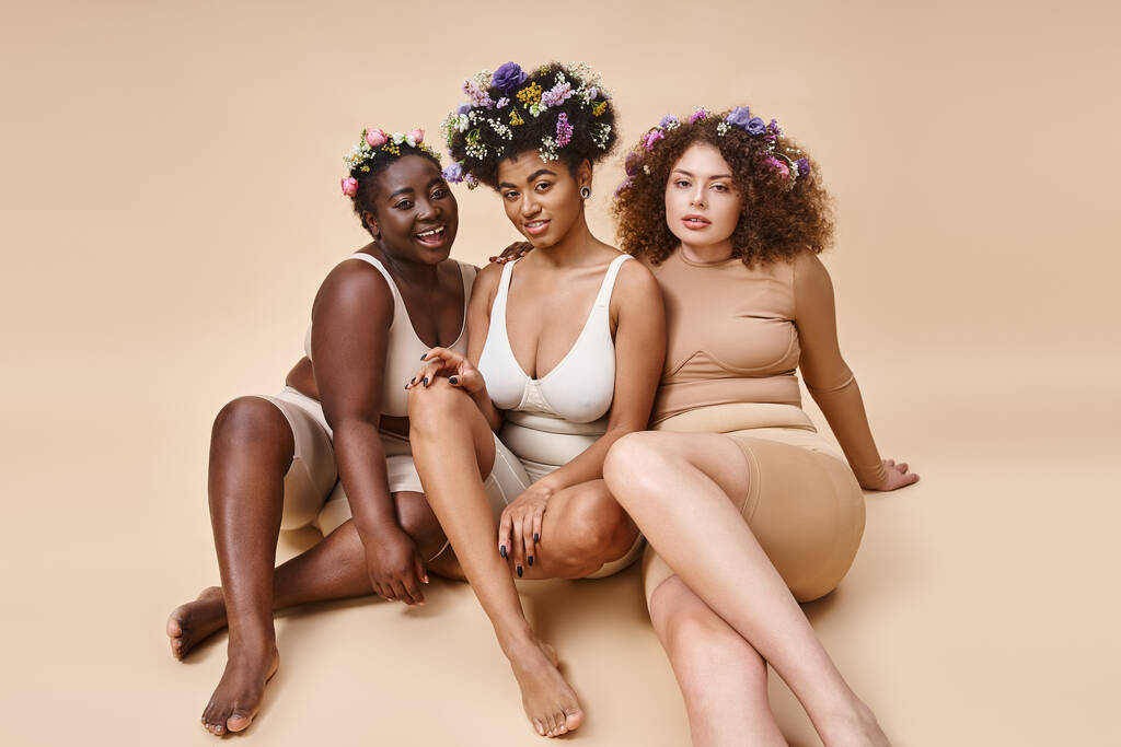diversas novias multiétnicas en lencería con flores en el pelo sentado en beige, además de belleza de tamaño - Foto, imagen