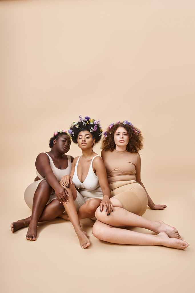 corps positif copines multiethniques en lingerie avec des fleurs dans les cheveux assis sur fond beige - Photo, image