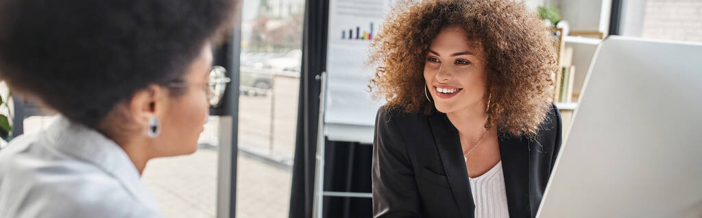 glückliche Geschäftsfrau im Gespräch mit afrikanisch-amerikanischer Kollegin mit Notizbuch in modernem Büro, Banner - Foto, Bild