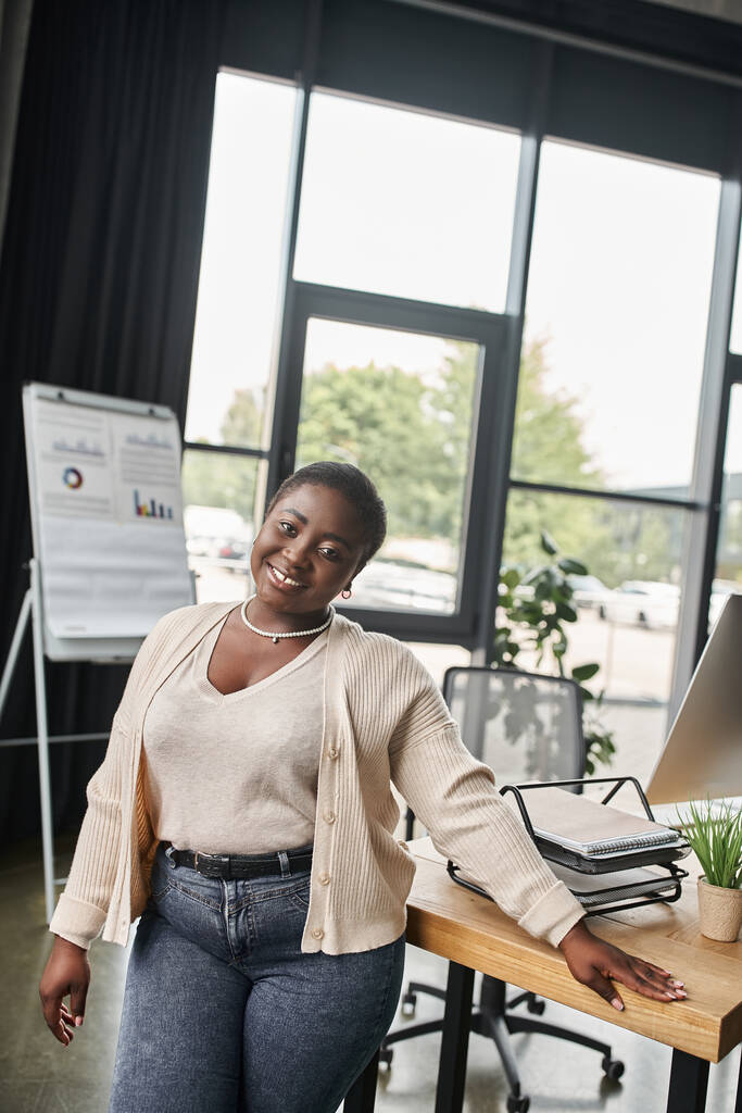 alegre africano americano curvilínea mujer de negocios mirando cámara cerca lugar de trabajo en moderno oficina - Foto, imagen