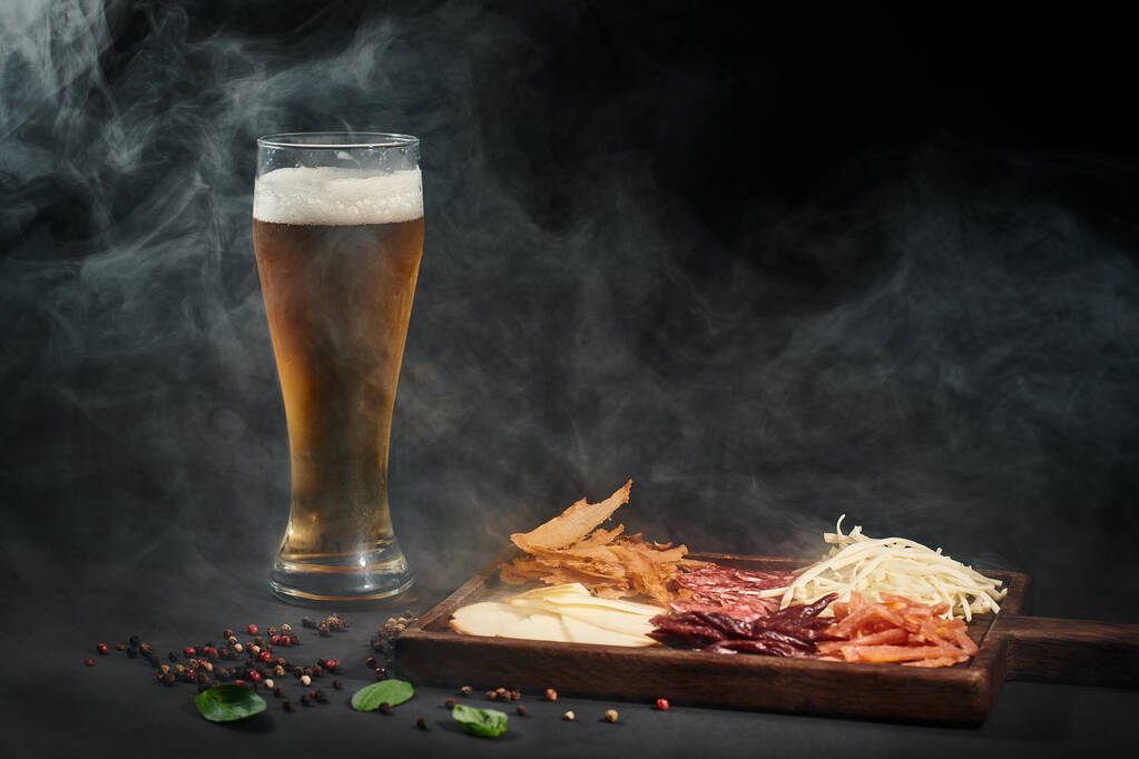 стакан пива рядом с доской для гурманов с сыром, сушеной говядиной и салями на черном фоне - Фото, изображение