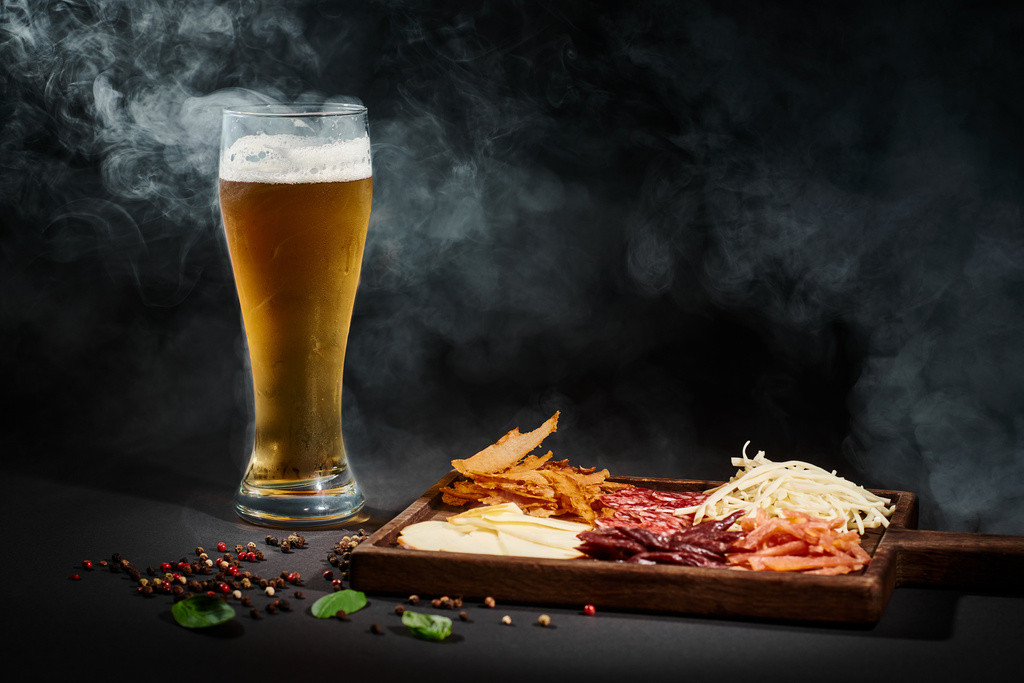 vaso de cerveza artesanal cerca del tablero de charcutería con selección de queso, carne seca y salami en negro - Foto, Imagen