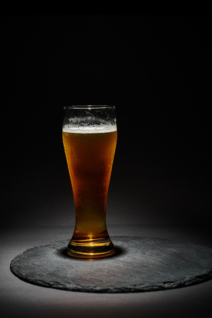 glass of craft beer or lager with foam in chilled glass on slate coaster on black background - Photo, Image
