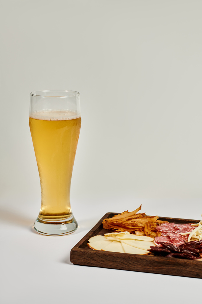 стакан ремесленного пива возле доски с сыром, сушеная говядина и салями на сером - Фото, изображение