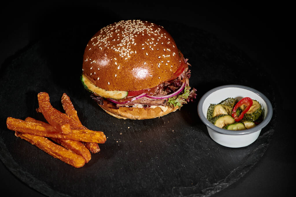 hambúrguer saboroso com carne dentro de pão de gergelim perto de picles como prato lateral no fundo preto - Foto, Imagem