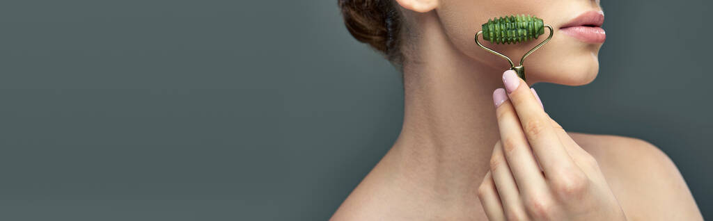 молода жінка з ідеальним обличчям для масажу шкіри з нефритом на сірому фоні, банер для догляду за шкірою - Фото, зображення