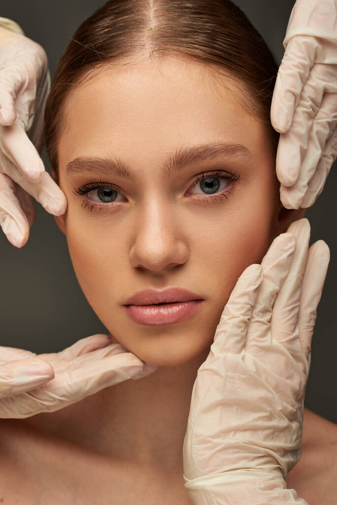 primer plano de los esteticistas en guantes médicos examinar la cara de paciente joven bonita sobre fondo gris - Foto, Imagen