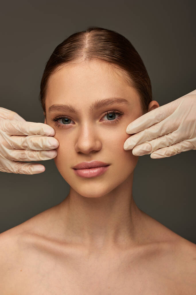 Tıbbi eldivenli estetisyenler gri arka planda güzel bir kadının yüzüne dokunuyor, dermatoloji konsepti. - Fotoğraf, Görsel