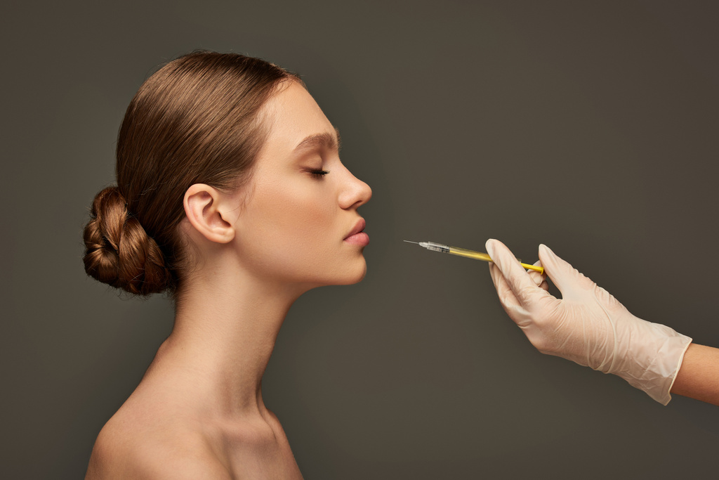 kozmetikus orvosi kesztyű tartó fecskendő közelében fiatal nő szürke háttér, oldalnézet - Fotó, kép