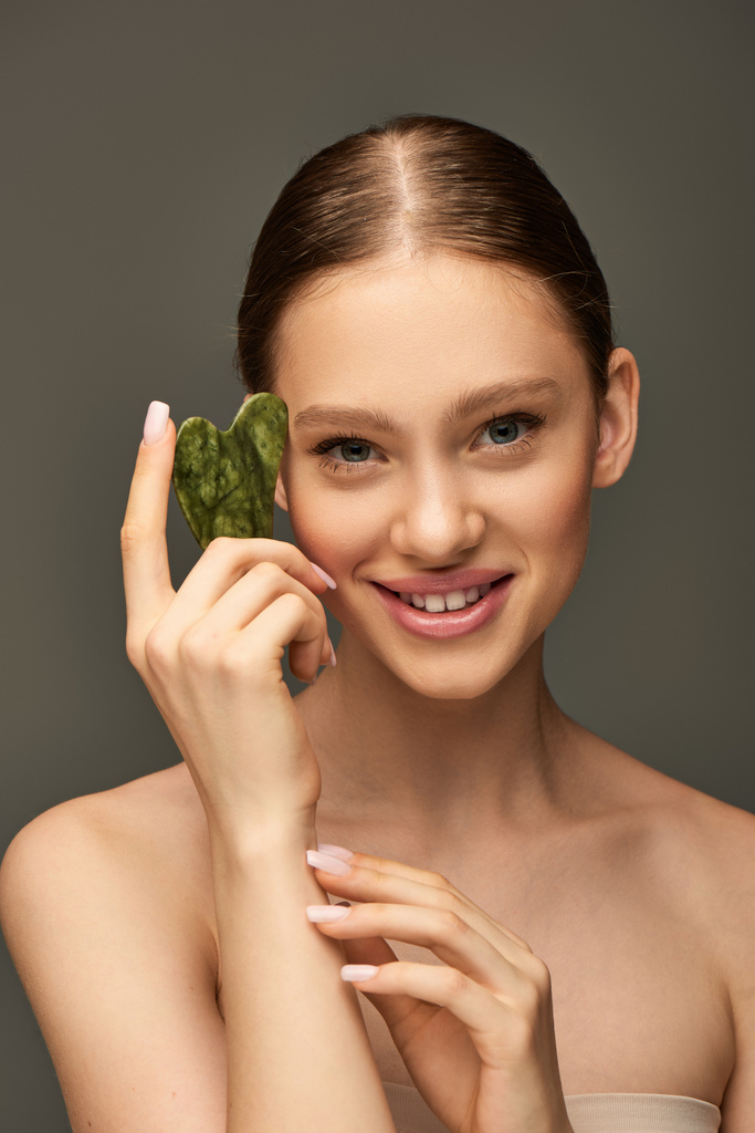 feliz joven mujer sonriendo y sosteniendo jade gua sha piedra sobre fondo gris, concepto de contorno facial - Foto, imagen