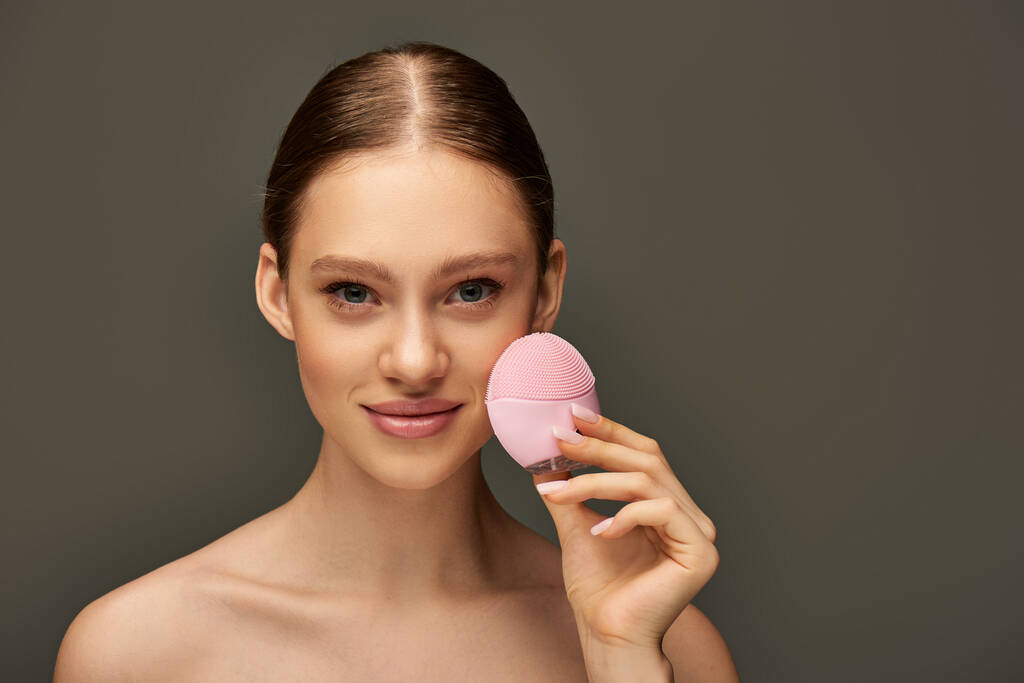 jonge vrouw met draagbare reinigen borstel op grijze achtergrond, schoonheid gadget en huidverzorging concept - Foto, afbeelding