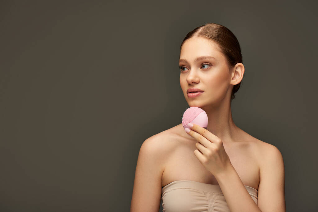 donna attraente che tiene spazzola detergente su sfondo grigio, gadget di bellezza e concetto di cura della pelle - Foto, immagini