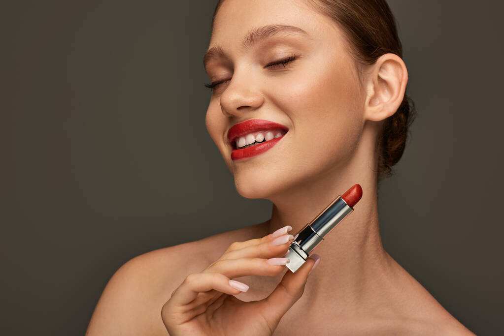 glückliche junge Frau mit roten Lippen, Lippenstift in der Hand und Lächeln auf grauem Hintergrund, Feiertags-Make-up - Foto, Bild