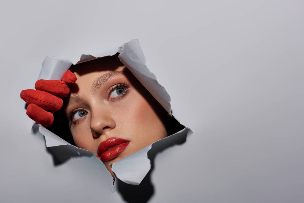 ελκυστική νεαρή γυναίκα με μπλε μάτια και κόκκινο κραγιόν κρυφοκοιτάζει μέσα από την τρύπα σε σχισμένο γκρι χαρτί - Φωτογραφία, εικόνα