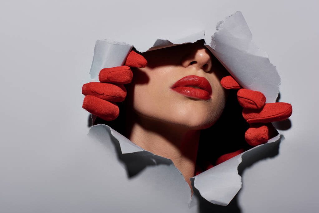vista recortada de mujer joven con labios rojos en guantes rasgando fondo gris y haciendo un agujero - Foto, imagen