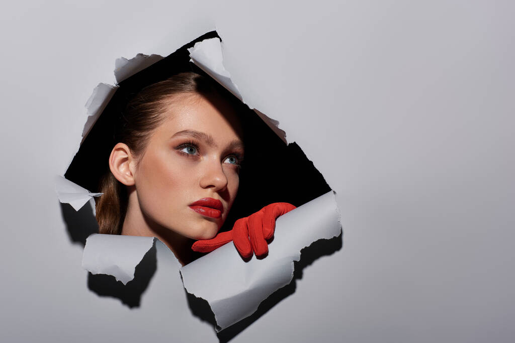 pretty young woman with blue eyes and red lipstick peeking through hole in ripped grey paper - Photo, Image