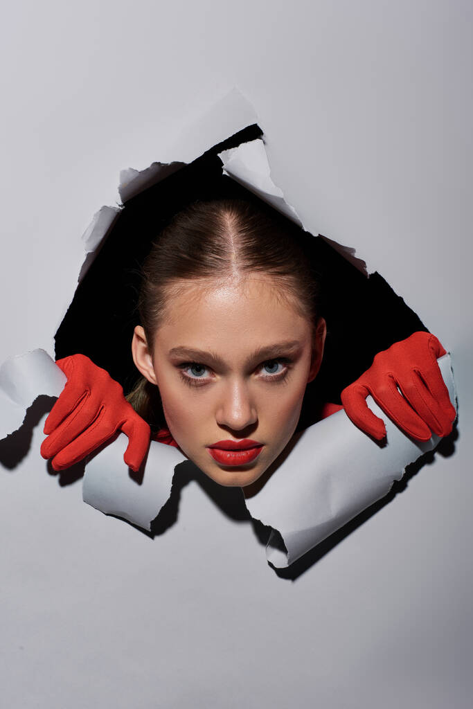 młoda kobieta z czerwoną szminką i rękawiczkami zaglądająca przez dziurę w szarym papierze, odważny makijaż - Zdjęcie, obraz