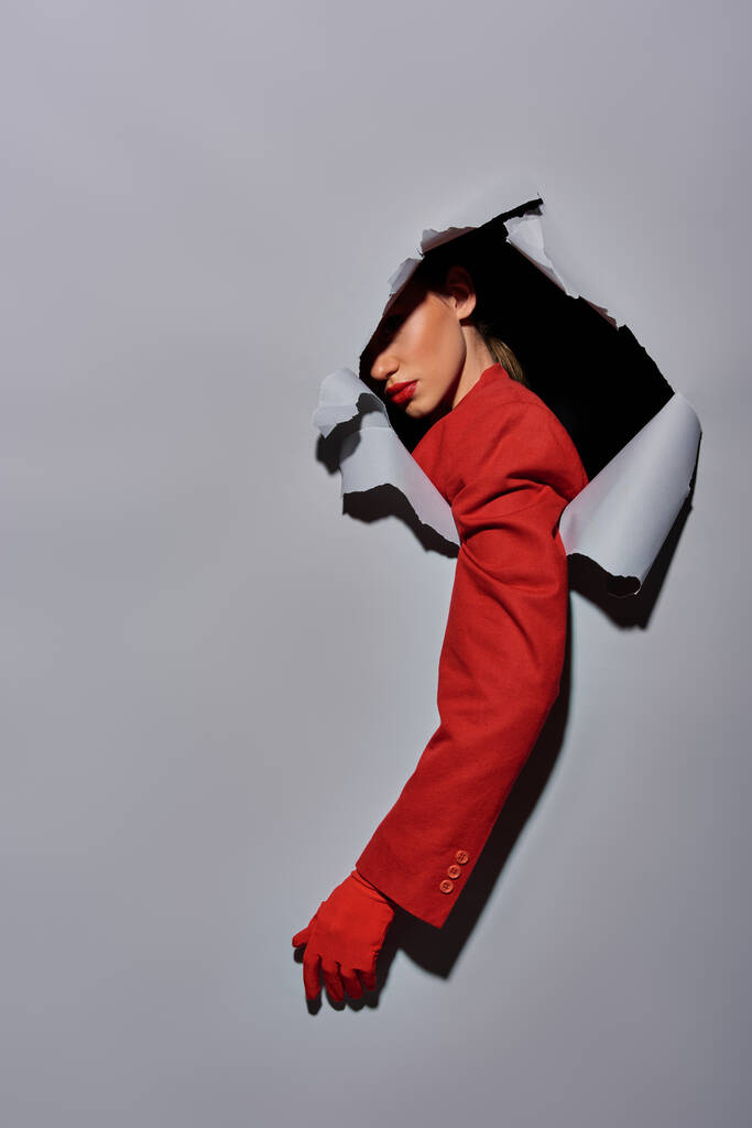 bijgesneden schot van vrouw met rode mouw en handschoen breken al gat in grijze achtergrond, conceptueel - Foto, afbeelding