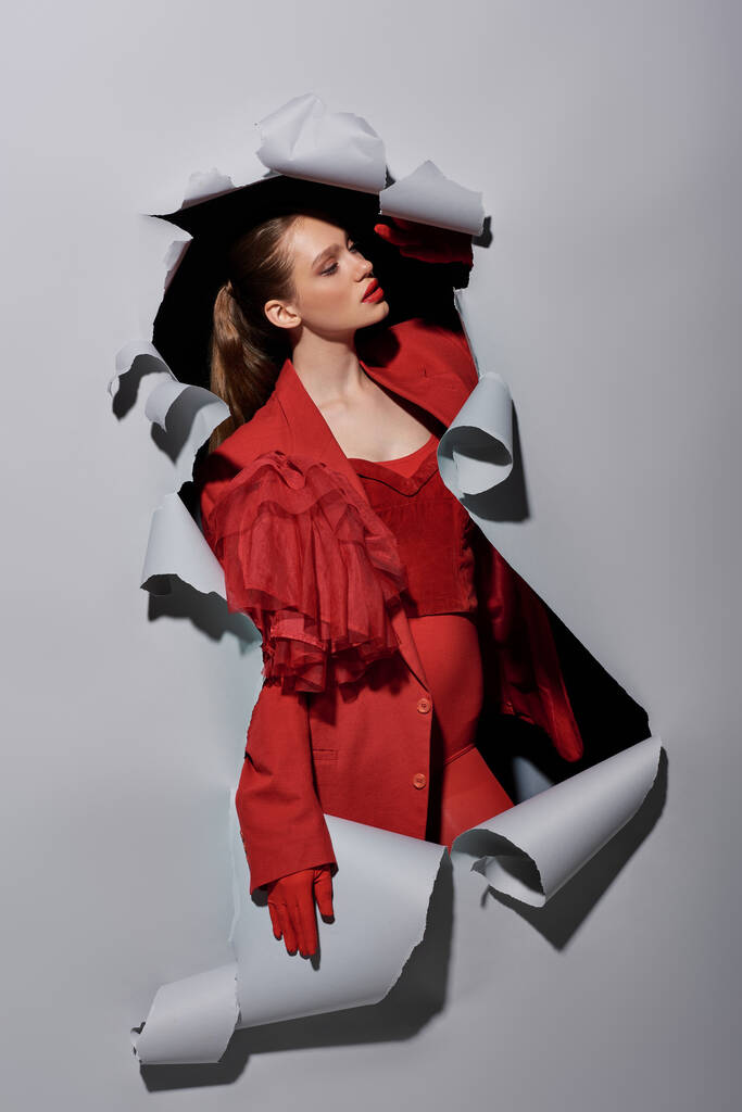 stylish woman in red blazer and gloves breaking though hole in torn grey background, conceptual - Photo, Image