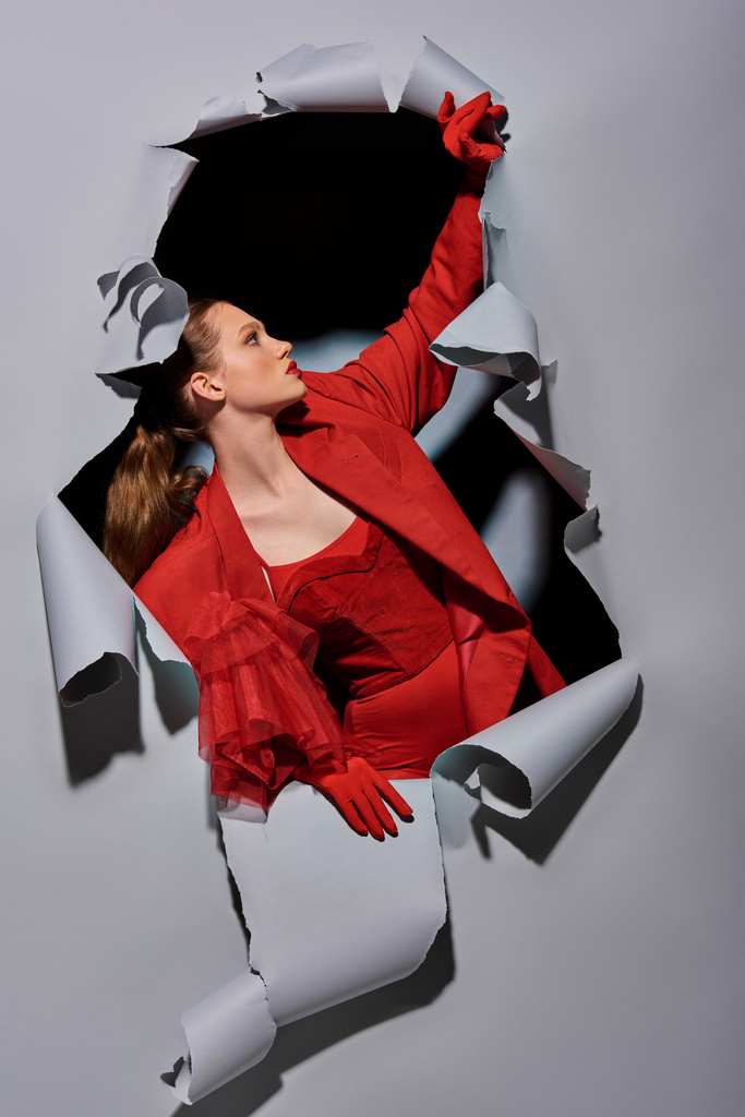 穴が付いている灰色の背景を壊す大胆な構造の赤い服のスタイリッシュな若い女性 - 写真・画像