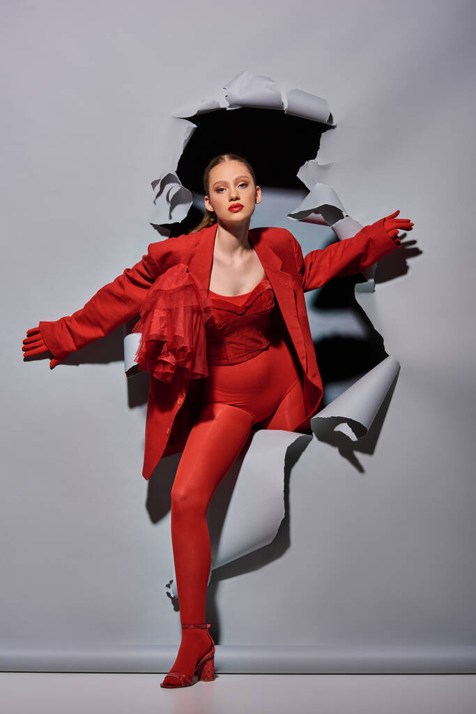 модная молодая женщина в красной одежде с смелым макияжем прорываясь через серый фон с отверстием - Фото, изображение