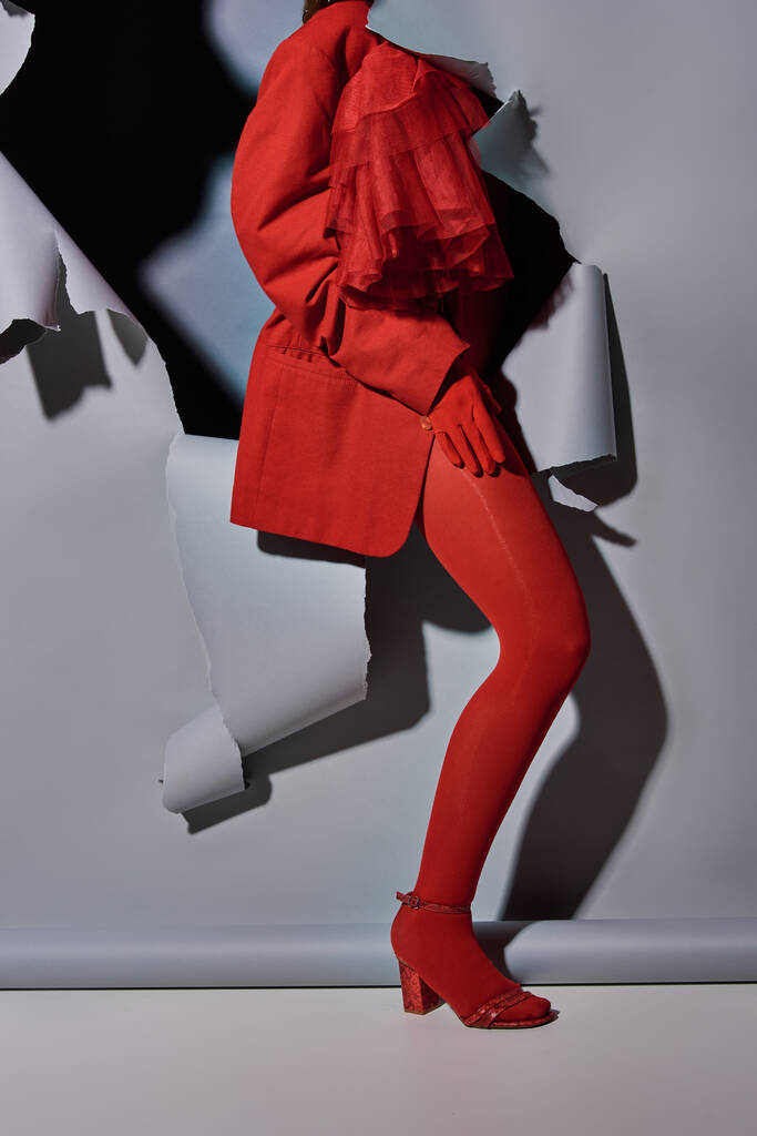 bijgesneden weergave van modieuze jonge vrouw in rode outfit doorbreken van grijze achtergrond met gat - Foto, afbeelding