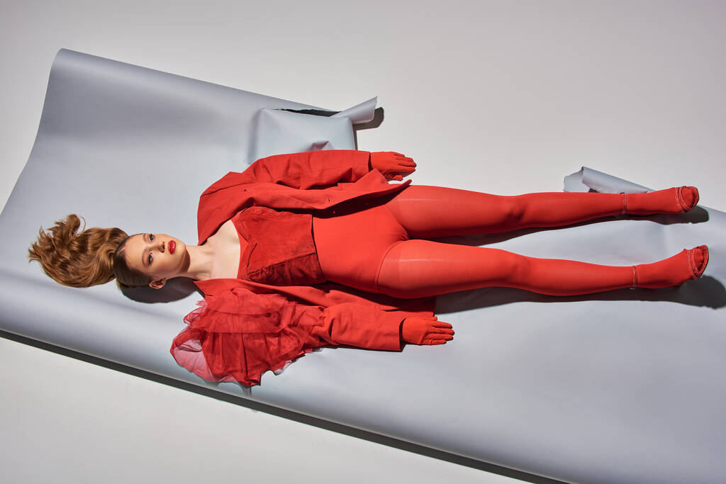 πάνω όψη του νεαρού μοντέλου με κόκκινη στολή ξαπλωμένη σε γκρι φόντο, γυναίκα που ενεργεί σαν κούκλα - Φωτογραφία, εικόνα