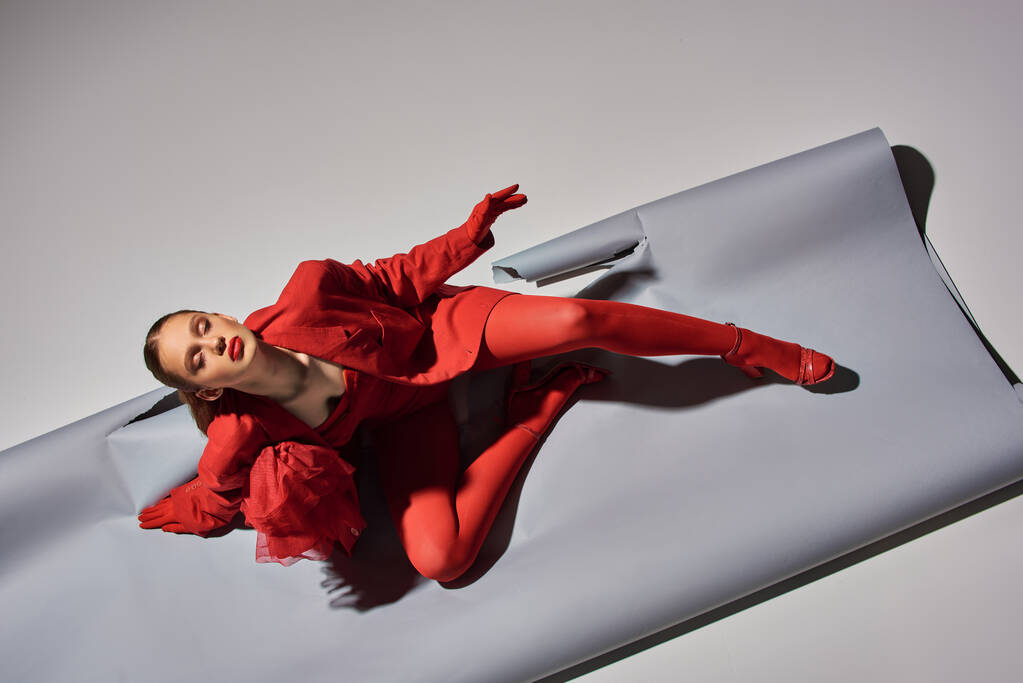 horní pohled na mladý model v červeném oblečení s punčocháče a vysoké podpatky sedí na šedém pozadí, móda - Fotografie, Obrázek