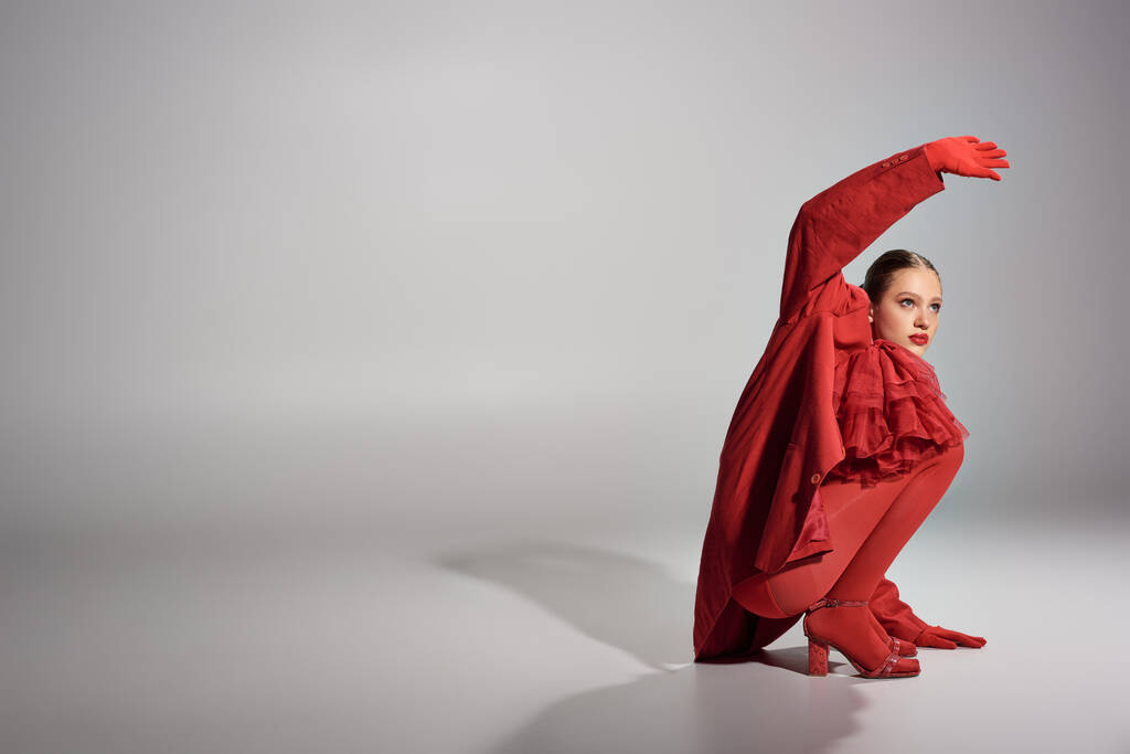 alta moda modelo jovem em roupa elegante vermelho posando com mão levantada no fundo cinza - Foto, Imagem