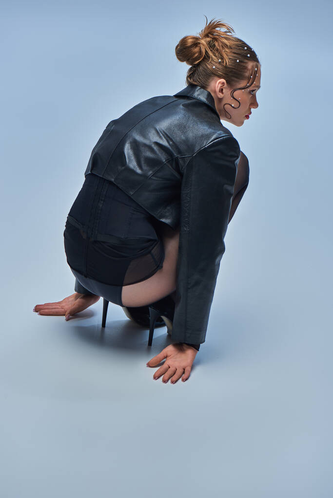 junges trendiges Model in Lederjacke sitzt in schwarzen Strumpfhosen und High Heels vor grauem Hintergrund - Foto, Bild