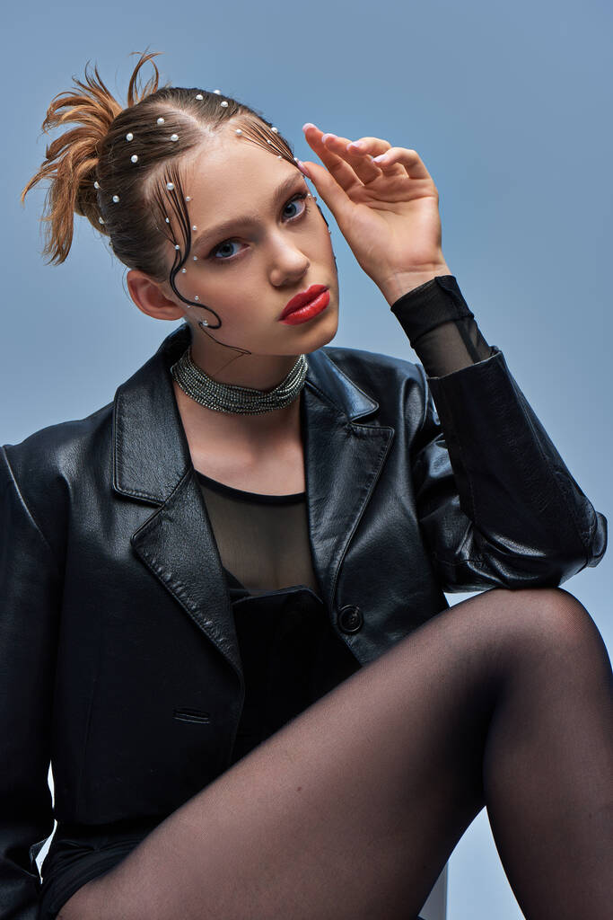 young trendy model in leather jacket and black tights looking at camera on grey backdrop, fashion - Photo, Image