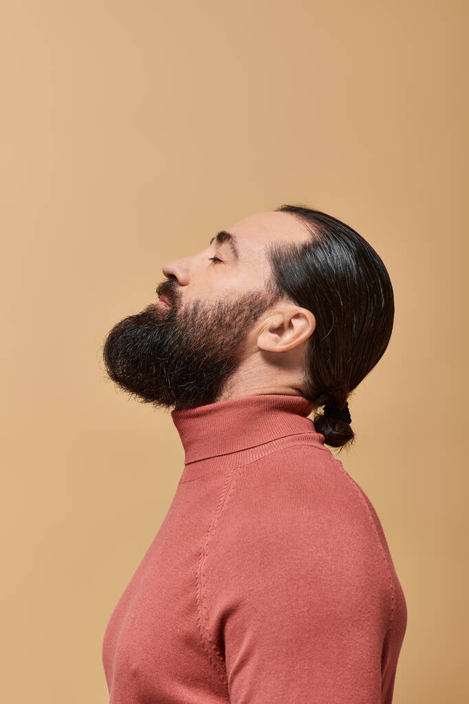 vue latérale du bel homme à la barbe posant en pull col roulé rose sur fond beige - Photo, image