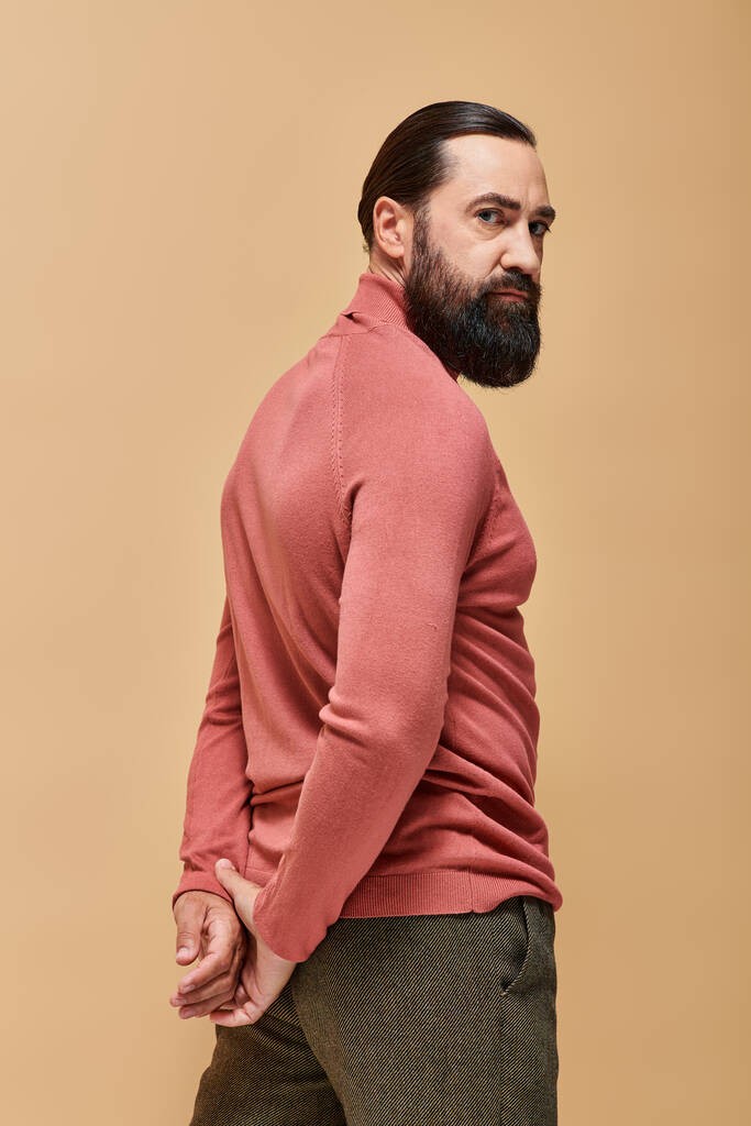 przystojny i poważny mężczyzna z brodą pozujący w różowym swetrze na beżowym tle, portret - Zdjęcie, obraz