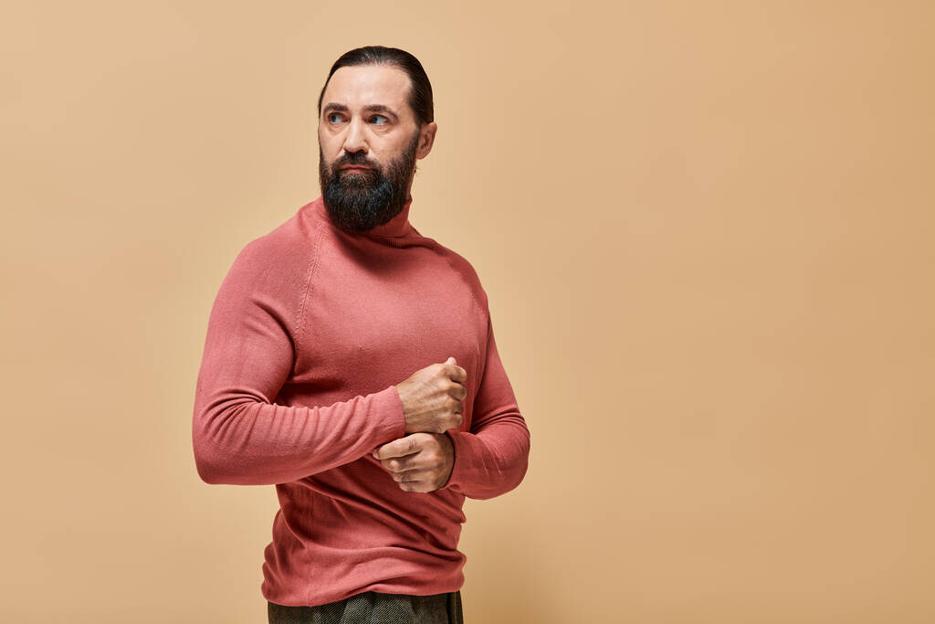 красивый и серьезный человек с бородой позирует в розовой водолазке свитер на бежевом фоне, портрет - Фото, изображение