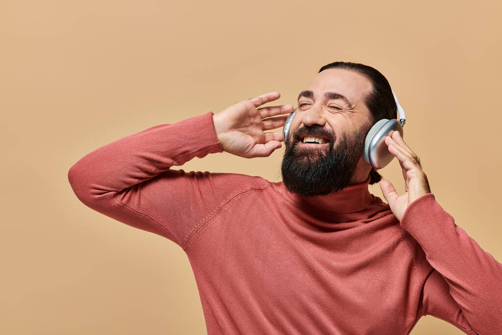 bärtiger positiver Mann im Rollkragenpullover, der Musik in drahtlosen Kopfhörern hört, beige Hintergrund - Foto, Bild